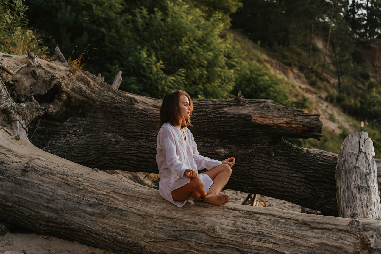 Woman in white shirt meditates on a large log in a forest setting during the day.